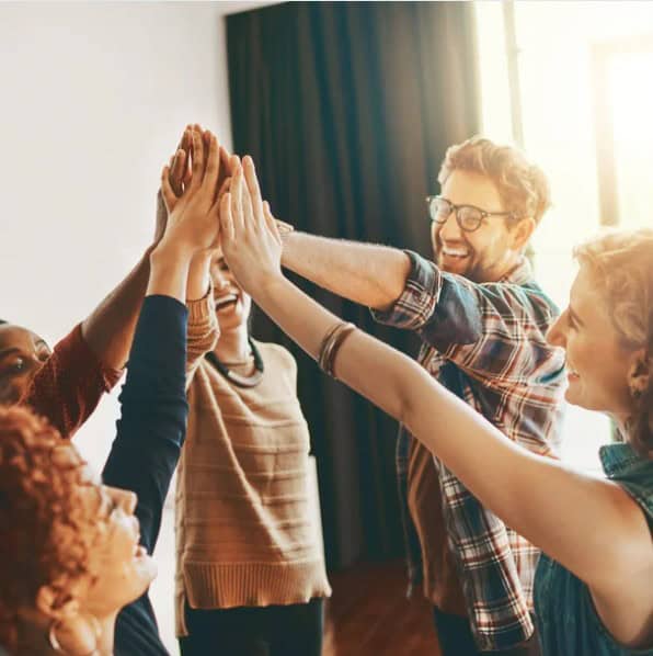 A group of people stand in a circle indoors, smiling and joining hands in a high-five—a moment inspired by professional development coaching.