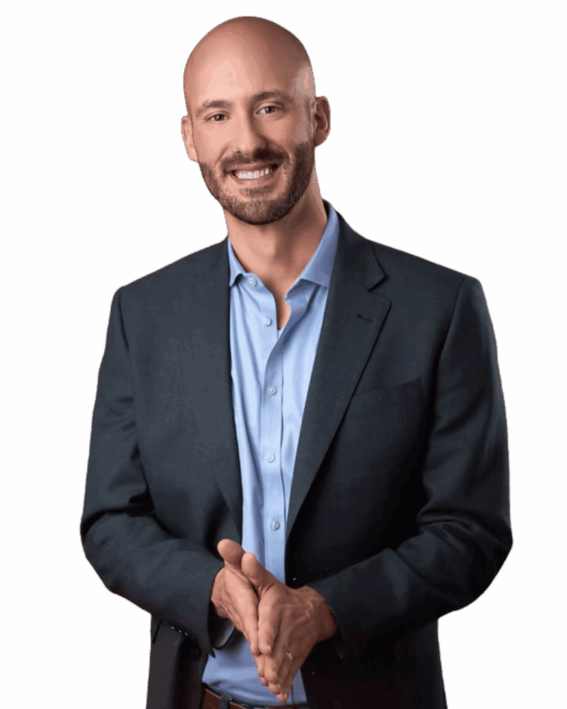 A man with a shaved head and beard smiles, wearing a dark suit jacket over an open-collared light blue shirt.